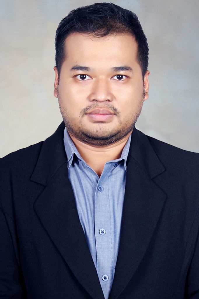 Foto resmi seorang pria mengenakan jas hitam dan kemeja biru, dengan ekspresi serius dan latar belakang berwarna netral.